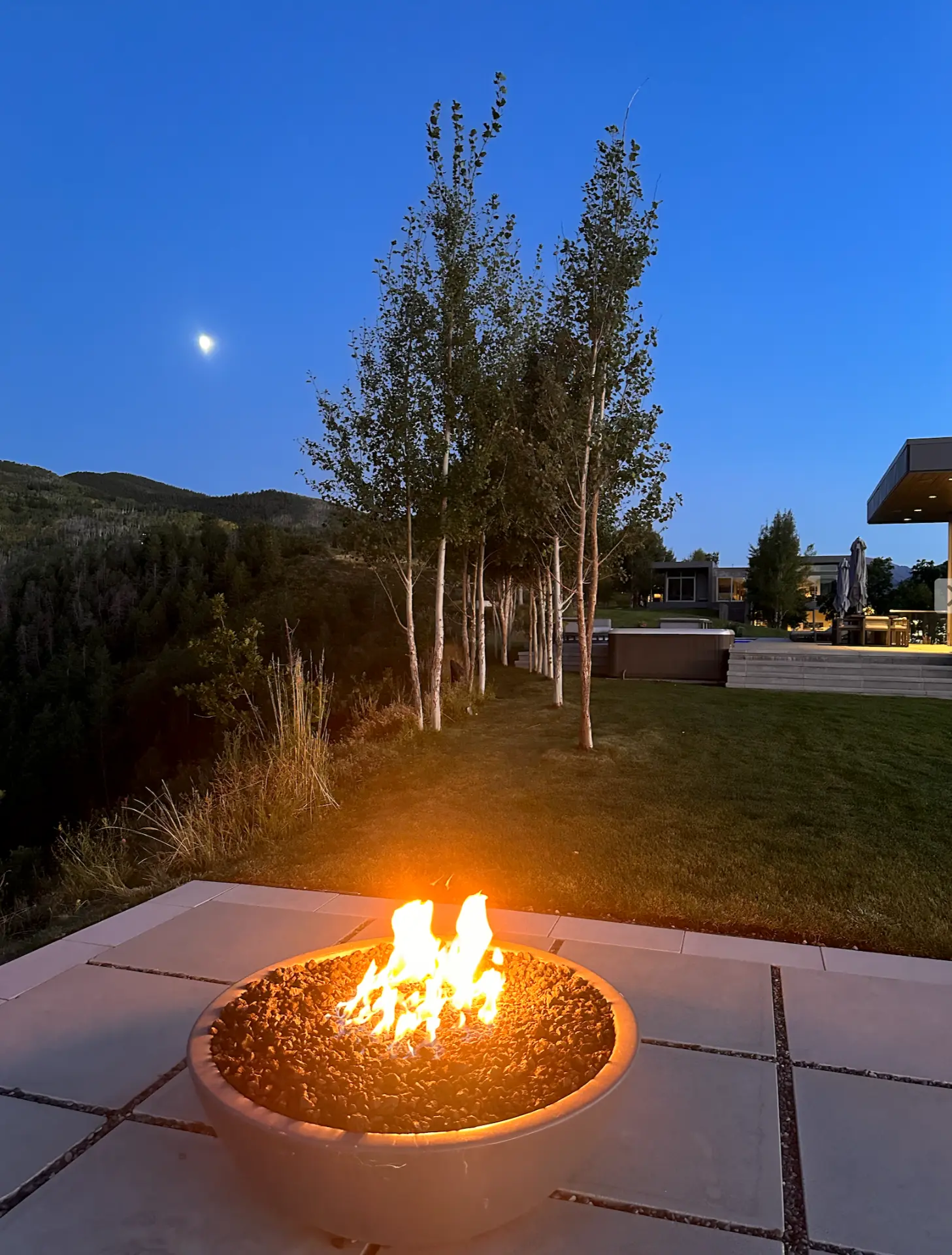 Firepit terrace at twilight with a modern mountain residence and moonlit ridge