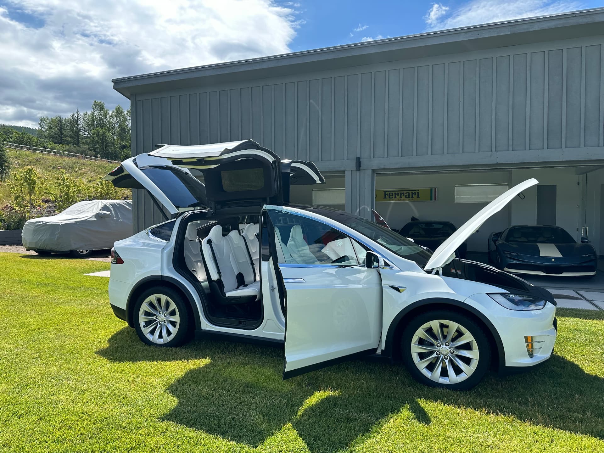 A white Tesla Model X with falcon doors open at a coach barn with covered collector cars